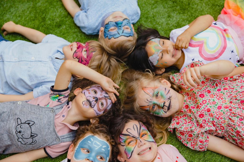 Group of children enjoying an outdoor party with colorful face paint, lying on grass.